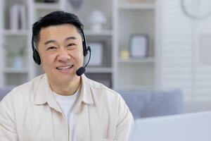 Friendly Asian male professional using a headset in a modern home office setting, looking cheerful and attentive while engaging in customer service or telecommuting. photo