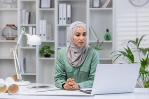 Woman in hijab designer working in home office, architect using laptop at work, studying technical assignment online, serious and concentrated reading. photo