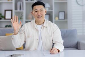 Cheerful Asian senior man sits at his desk at home, happily writing notes while greeting with a wave, adding a friendly and inviting feel. photo
