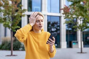 A middle-aged woman appears distressed and worried while she holds a smartphone on a sunny day with an urban background. Her expression conveys shock and confusion. photo