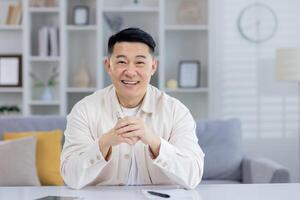un alegre asiático hombre sentado cómodamente a hogar, exudando felicidad y confianza con un brillante, acogedor sonrisa. moderno vivo habitación ajuste. foto