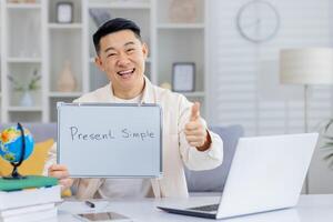 Asian teacher happily giving a thumbs up while teaching the present simple tense online. Bright home office setting with laptop, books, and a globe, creating an engaging and educational environment. photo