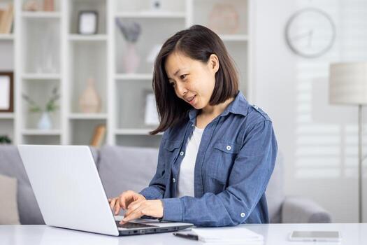 un profesional asiático mujer es absorto en trabajo en un computadora portátil, sentado en un bien iluminado y pulcramente arreglado vivo habitación reflejar comodidad y productividad ese es ideal para remoto trabajo o personal proyectos foto