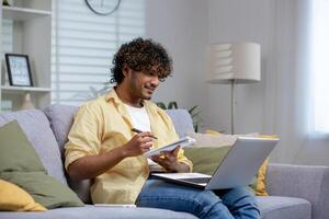 A young adult sits comfortably on a sofa, working on a laptop while taking notes in a notebook, depicting a productive work-from-home setting. photo