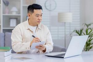 Man working from home, sitting at a desk with a laptop and taking notes in a relaxed and modern living space, demonstrating productivity and concentration. photo