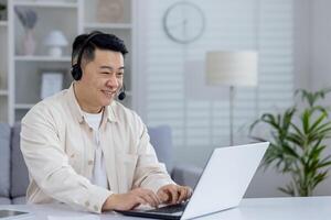 Cheerful Asian man wearing a headset and working on his laptop from a well-lit home office. He appears engaged and happy with his task. photo