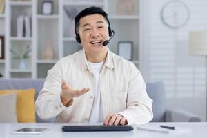 Cheerful Asian male in a beige shirt using a headset in a home office, looking at camera with a smile, possibly in a customer service call. photo