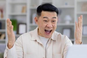 A middle-aged Asian man shows great excitement and joy while working at his computer in a modern home office setting. His exhilarated expression suggests a moment of triumph. photo