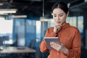 A focused Asian businesswoman with a serious expression is thinking deeply while using a digital tablet in a modern office setting. photo