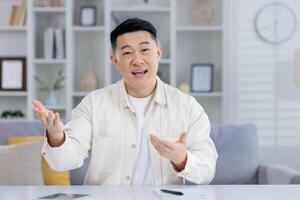Cheerful Asian man engaging in a casual conversation at home, looking at the camera and talking with expressive hand gestures in a modern living room setting. photo
