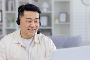 Cheerful Asian man wearing a headset and smiling while using a laptop in a bright home office. Workspace concept with a focus on remote work and communication. photo