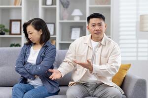 Asian couple sitting on a sofa in living room. The woman looks upset and withdrawn, while the man gestures as if trying to explain something, conflicts in relationships. photo