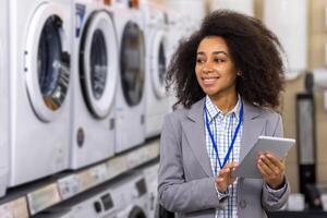 In an appliance store, a happy woman with curly hair smiles holding a tablet. She is comparing options in front of washing machines and dryers, possibly making a purchase or gathering information for buying photo