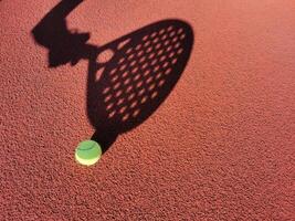 High angle of yellow ball placed on court and covered with shadow of crop person holding padel racket photo