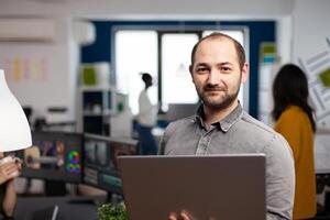 editor worker standing in front of camera smiling working in creative agency office holding laptop. Man videographer works in multimedia studio production editing in modern workplace. photo
