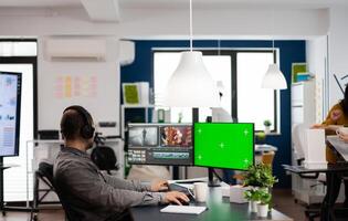 Man videographer with headset editing movie in post production software working at greenscreen, chroma key isolated display of computer. editor processing audio film montage in start up office photo