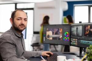 Videographer looking at camera smiling working in creative startup workplace using editing post production software on professional pc with two monitors processing film montage. photo