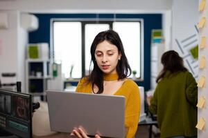 Focused entrepreneur standing in creative agency office holding laptop and typing project informations. Photographer graphic editor working in start up digital multimedia company, using software app photo