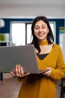 Woman retoucher looking at camera smiling working in creative media agency standing in multimedia company holding laptop. Photographer graphic editor working in start up office at digital assets. photo