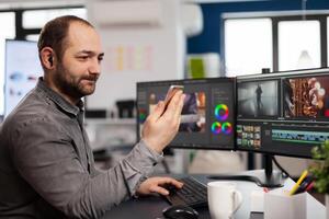editor talking on videocall with client holding smartphone editing movie sitting in creative agency. Videographer processing film montage in multimedia company with post production software photo