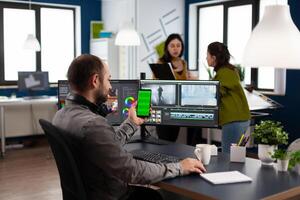 editor talking on call holding smartphone with green screen, chroma key mock up isolated display taking break from retouching movie sitting in start up office. Team working in background photo