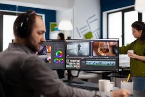 Videographer with headset working with footage and sound on pc with two displays, taking notes while editing customer . Man processing film montage sitting in creative startup agency office. photo