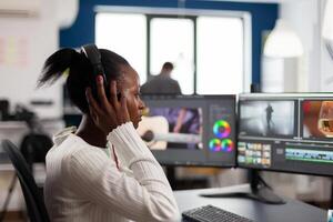 African editor with headset listening music wihile editing footage and sound sitting in creative agency. Videographer in digital multimedia company processing movie with post production software photo