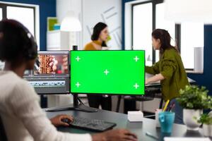 African woman videographer with headset processing film montage working at greenscreen, chroma key isolated display of computer. editor editing movie in post production software in agency office photo