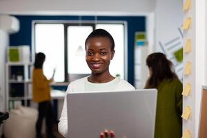 Close up of african woman looking at camera smiling standing in start up creative agency office holding laptop, typing on it. Black videographer editing project using post production software photo
