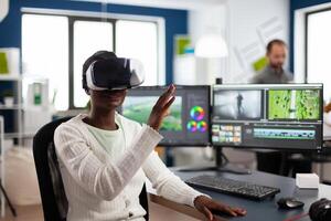 African editor experiencing VR goggles, gesturing, editing film montage working with footage and sound on computer with two displays. Videographer processing movie in creative agency photo