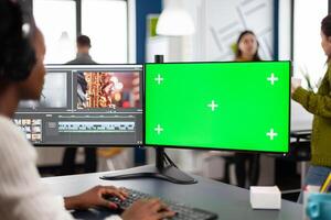 African editor with headset editing footage using pc with greenscreen, chroma key isolated display sitting in creative agency. Videographer in digital multimedia company processing movie photo
