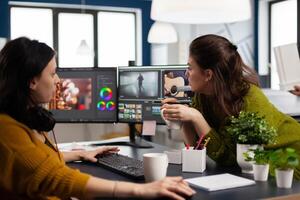 Coworkers editors discussing in front of pc working for client footage using post production software and two monitors. Creative People processing film montage in digital multimedia company photo