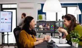 Team of videographers taking work break discussing and laughing, drinking coffee working in editing app at pc sitting in digital multimedia company. Creative women prosessing film montage photo