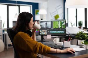 Professional colorist working in footage using post production software, drinking coffee. Videographer editing audio film montage at pc with two monitors, sitting on desk in creative agency photo