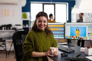 Woman retoucher looking at camera smiling sitting in creative media agency retouching client photos on PC with two displays. Photographer graphic editor working in start up office with digital assets