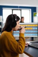 Photo editor with headphones retouching image using stylus pen, drinking coffee, working on PC with two displays. Woman retoucher editing in software app with graphic tablet sitting in creative agency