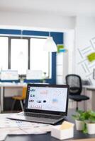Close up of laptop in start up company office with charts and statistics. Brainstorm area in business center with nobody in it, shot of empty room with modern furniture and blue wall. photo