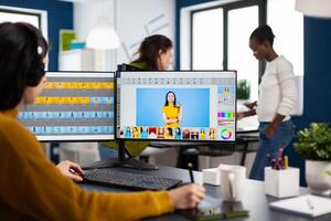 Woman retoucher with headset retouching photos using stylus pen drawing on graphic tablet, working on PC with two displays. Photo editor working in software app sitting in creative agency office