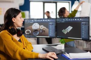 Industrial female engineer looking at personal computer with dual monitors setup, screens showing CAD software with 3D prototype of gears metalic mechanical piece. Employee working in creative office photo