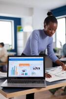 Laptop with financial charts on office of start up company and african woman reading statistics. Diverse team of business people analyzing company financial reports from computer. Successful corporate professional entrepreneur online internet statistics photo