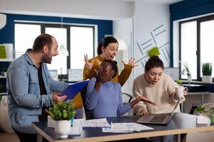 Diverse business people screaming after corporate achievement in startup office. Multiethnic coworkers business team with laptop and papers excited about project. photo
