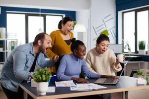 Excited diverse coworkes in statup company office celebrate big sales triumph. Happy multiethnic colleagues business team with laptop and papers excited about project. photo