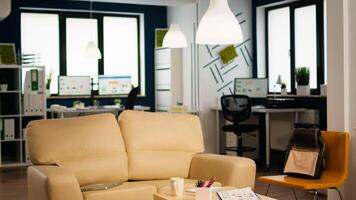 Interior of cozy light company room with small table and confortable couch, modern stylish chairs and desktop computer, all ready for employees. Empty spacious office of creative working space. photo