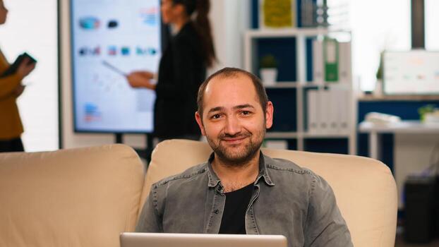 gerente sentado en sofá en frente de cámara sonriente mientras multiétnico equipo trabajando en antecedentes con financiero informes. diverso compañeros de trabajo hablando acerca de puesta en marcha financiero empresa en moderno oficina. foto