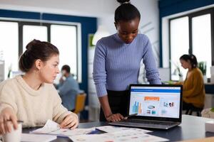 Business manager leadership briefed about financial charts by african employee looking at laptop display. Multiethnic employees gathered in co-working space. photo