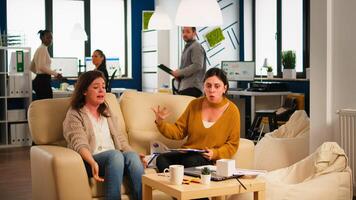 Stressed busineswomen arguing, yealing at each other during work hours sitting on the couch, while diverse colleagues working scared in background. Women throwing papers, conflicts at work concept photo