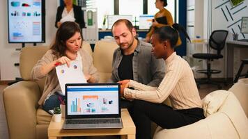 Diverse colleagues employees cooperating in office discussing at workplace, analysing graphs sitting on couch. Business coworkers planning start up business project, brainstorming in corporate office photo
