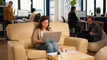 Employee holding laptop sitting in relax zone on confortable couch typing on pc smiling while diverse colleagues working in background. Multiethnic coworkers planning new financial project in company photo