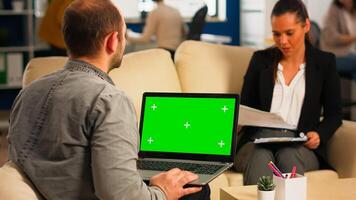 Back view of business man sitting on couch using laptop with green screen talking with colleague while team working on background. Multiethnic coworkers planning project on chroma key display photo