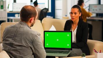 Business team sitting on couch analysing financial statistics, holding laptop with green screen while diverse team working on background. Multiethnic coworkers planning project on chroma key display photo